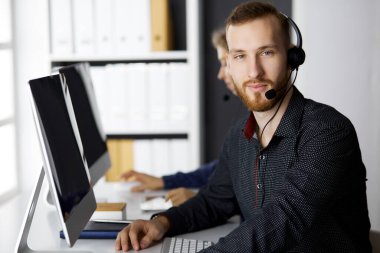 Modern ofiste otururken kulaklıkla konuşan sakallı iş adamı. Çağrı merkezinde çeşitli insanlar var. Tele pazarlama ve müşteri hizmetleri