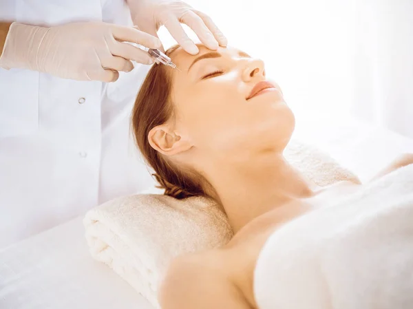 Beautiful woman receiving beauty injections with closed eyes in sunny medical center. Beautician doctor hands doing beauty procedure to female face with syringe. Cosmetic medicine and surgery concept