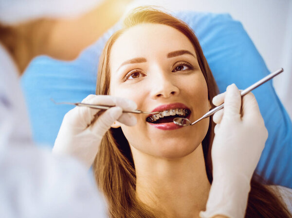 Smiling young woman with orthodontic brackets examined by dentist in sunny dental clinic. Healthy teeth and medicine concept