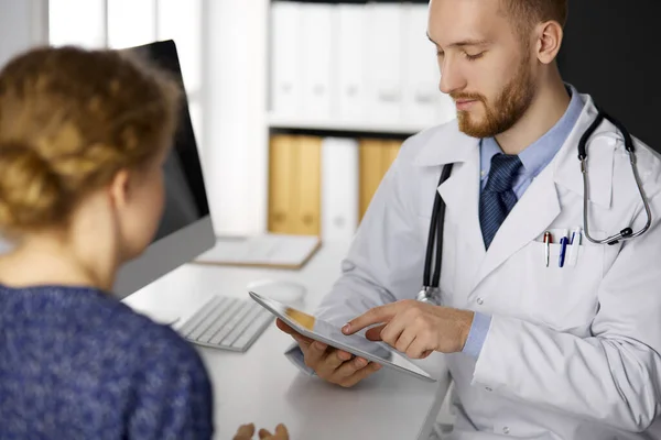Amistoso médico de barba roja y mujer paciente discutiendo el examen de salud actual mientras está sentado y el uso de tableta en la clínica — Foto de Stock