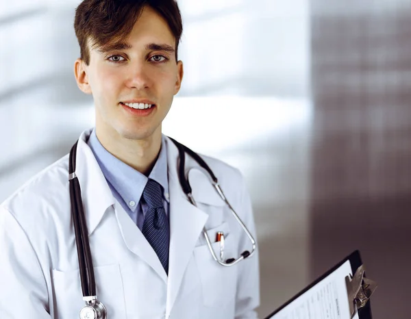 Barátságos fiatal férfi orvos áll írótáblával a modern klinikán. Orvosi fogalom — Stock Fotó