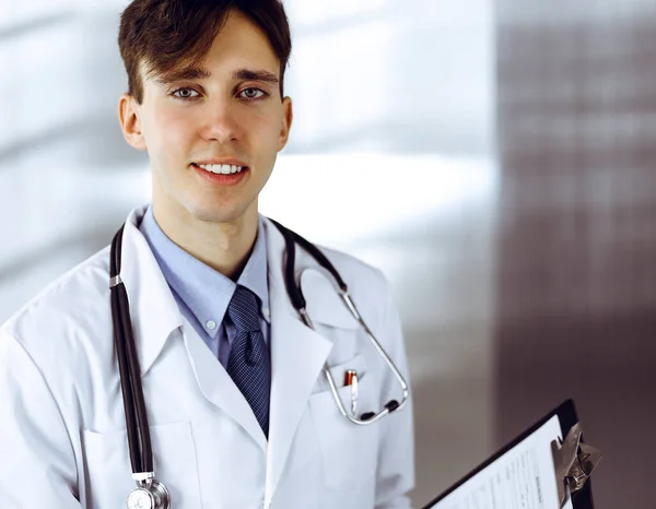 Freundlicher junger Arzt mit Klemmbrett in moderner Klinik. Medizinkonzept Stockbild