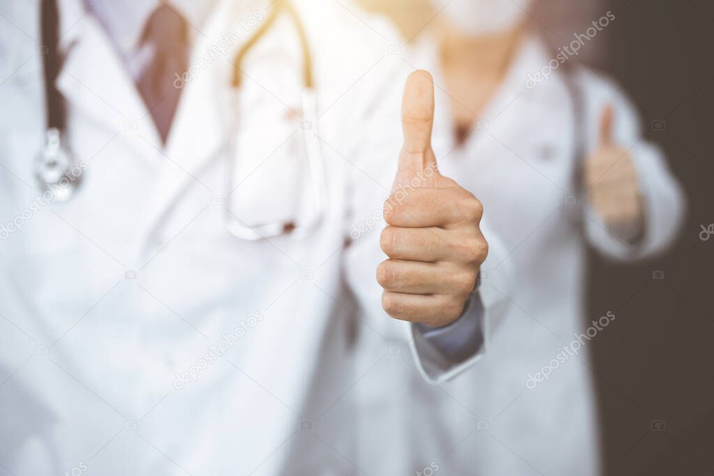 Unknown man-doctor and woman standing straight as a team with thumbs up in sunny clinic. Medicine concept during Coronavirus pandemic. Covid 2019