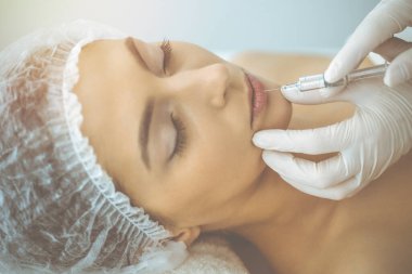 Güzellik uzmanı güneşli klinikte genç esmer kadının yüzüne şırıngayla güzellik prosedürü uyguluyor. Kozmetik ilaç ve ameliyat, güzellik iğneleri.