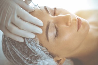 Güzellik uzmanı güneşli klinikte genç esmer kadının yüzüne şırıngayla güzellik prosedürü uyguluyor. Kozmetik ilaç ve ameliyat, güzellik iğneleri.