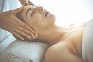 Güzel esmer kadın güneşli spa merkezinde gözleri kapalı yüz masajından hoşlanıyor. Rahatlatıcı tedavi ve kozmetik tıp kavramları