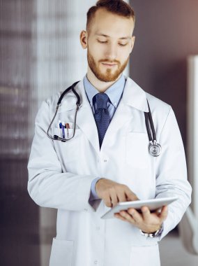 Dost canlısı sakallı doktor iş yerindeki güneşli klinikte tablet bilgisayar kullanıyor. Tıp konsepti