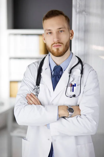 Vörös szakállú orvos áll egyenesen a klinikán, közel a munkahelyéhez. Orvosi portré. Gyógyszer, egészségügy — Stock Fotó