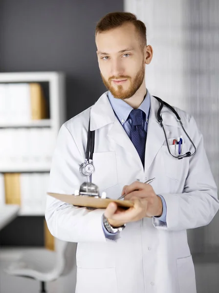 Friendly red-bearded doctor standing and writing with clipboard in clinic at his working place. Perfect medical service in hospital. Medicine, healthcare — Stock Photo, Image
