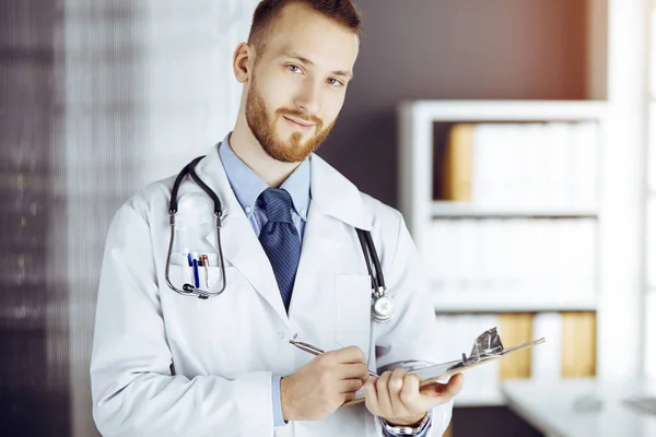 Φιλικός γενειοφόρος γιατρός στέκεται και γράφει με πρόχειρο στην ηλιόλουστη κλινική στο χώρο εργασίας του. Έννοια ιατρικής — Φωτογραφία Αρχείου