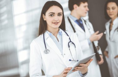 Gülümseyen zeki kadın-doktor, klinikte meslektaşlarıyla birlikte ayakta dururken elinde bir tablet bilgisayar tutuyor. Doktorlar iş başında. Hastanede mükemmel bir tıbbi hizmet.