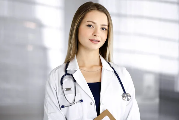 Giovane donna-medico ottimista tiene in mano una cartellina, mentre si trova in una clinica. Ritratto di medico donna amichevole con stetoscopio. Un servizio medico perfetto in ospedale. Medicina — Foto Stock