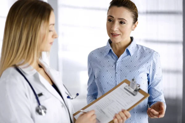 Jovem médica e sua paciente estão discutindo os pacientes exame de saúde atual, enquanto estão juntos em um consultório hospitalar. A médica está a escrever algumas marcas, usando uma prancheta. Perfeito. — Fotografia de Stock