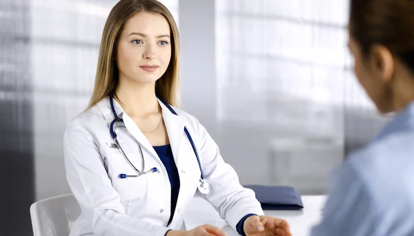 Νεαρή έξυπνη γυναίκα-γιατρός μιλάει με χαμόγελο στον ασθενή της, ενώ κάθονται μαζί στο ντουλάπι σε μια κλινική. Γυναίκα γιατρός με στηθοσκόπιο δίνει κάποιες συστάσεις σε ένα — Φωτογραφία Αρχείου