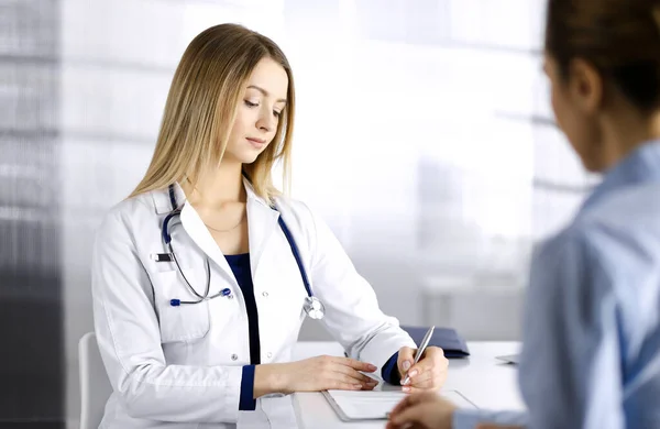 Ung intelligent kvinna-läkare lyssnar på hennes patienter allergier symptom, medan de sitter tillsammans i kabinettet på en klinik. Kvinnlig läkare, med ett stetoskop, skriver på — Stockfoto
