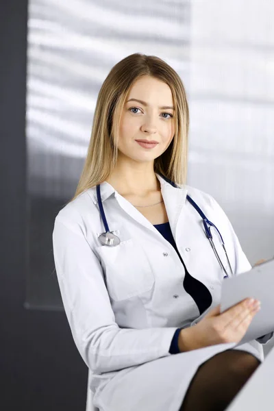 Jovem mulher-médico otimista está usando uma área de transferência, enquanto sentado na mesa em seu gabinete em uma clínica. Retrato de mulher médica amigável com um estetoscópio. Serviço médico perfeito em um — Fotografia de Stock