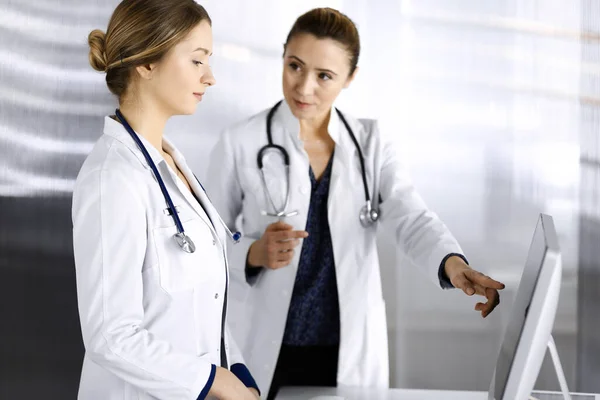 Two female physicians are discussing medical therapy, while standing at the table in a clinic office. Doctors use pc computer at work. Teamwork in medicine