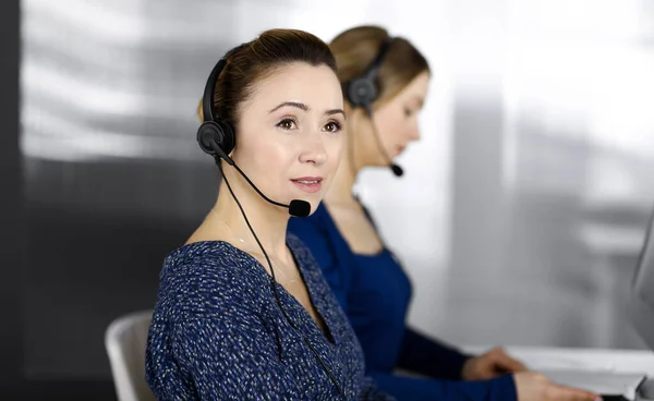 Two busineswomen have conversations with the clients by headsets, while sitting at the desk in a modern office. Diverse people group in a call center. Telemarketing and customer service