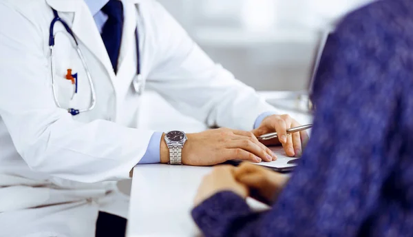Bilinmeyen genç bir hasta, hastane bürosundaki masada otururken, tıbbi testlerinin sonuçlarını bir doktorla tartışıyor. Doktor ilaç geçmişi kayıtlarını doldurmak için pano kullanıyor — Stok fotoğraf