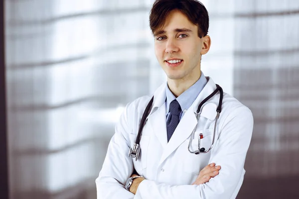 Allegro sorridente uomo-dottore in piedi con le braccia incrociate in clinica. Servizio medico perfetto con giovane medico intelligente in ospedale. Concetto di medicina — Foto Stock