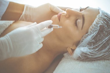 Güzellik uzmanı güneşli klinikte genç esmer kadının yüzüne şırıngayla güzellik prosedürü uyguluyor. Kozmetik ilaç ve ameliyat, güzellik iğneleri.