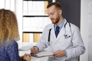 Dost canlısı kızıl sakallı doktor ve hasta kadın klinikte otururken sağlık muayenesini tartışıyorlar.