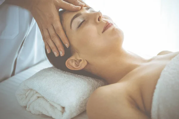 Piękna brunetka korzystająca z masażu twarzy z zamkniętymi oczami w słonecznym centrum spa. Relaksujące koncepcje leczenia i medycyny kosmetycznej — Zdjęcie stockowe