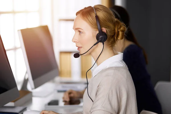 Femme d'affaires parlant par casque tout en étant assis dans un bureau ensoleillé. Centre d'appels et divers groupes de personnes en affaires — Photo