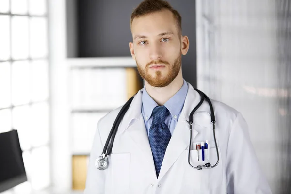 Rode baard arts staat recht in de kliniek in de buurt van zijn werkplek. Portret van een arts. Geneeskunde, gezondheidszorg — Stockfoto