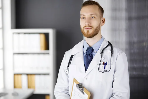 Red-bearded doctor standing straight in clinic near his working place. Portrait of physician. Medicine and healthcare concept