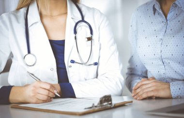 Bilinmeyen bir kadın, doktor ve hastası birlikte klinikteki dolapta otururken hastaların kan testlerini tartışıyorlar. Steteskoplu bir kadın doktor dizüstü bilgisayar kullanıyor.