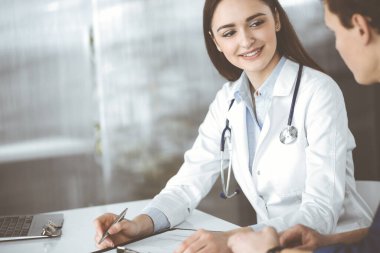 Dost canlısı genç bayan-doktor, birlikte bir dolapta masada otururken, hastasını dinliyor. Hekim elinde ilaç doldurmak için bir dosya tutuyor.