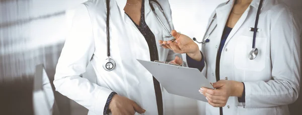 Dos médicas están discutiendo las pruebas médicas de sus pacientes, mientras están de pie en una clínica. Los médicos usan un portapapeles en el trabajo. Trabajo en equipo en medicina —  Fotos de Stock