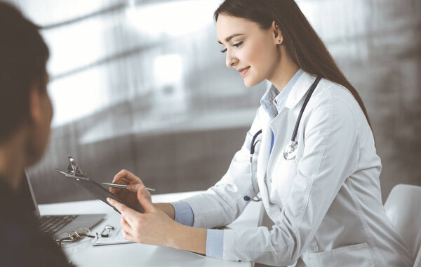 Friendly young woman-doctor is listening to her patient, while they are sitting together at the desk in a cabinet. Physician is holding a clipboard in her hands for filling up medication