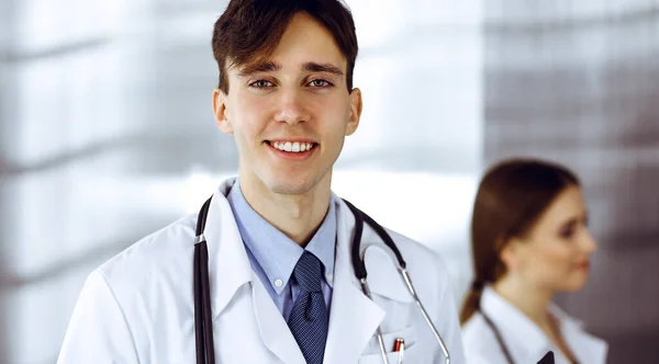 Vriendelijke jonge mannelijke arts staat met klembord in moderne kliniek. Vrouwelijke collega is bezig met het invullen van medicatiegegevens formulier op de achtergrond van de arts. Geneesmiddelenconcept — Stockfoto