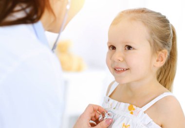 Doktor steteskopla küçük bir kızı muayene ediyor. Her zamanki tıbbi muayenede gülümseyen çocuk hasta. Tıp ve sağlık hizmetleri kavramları