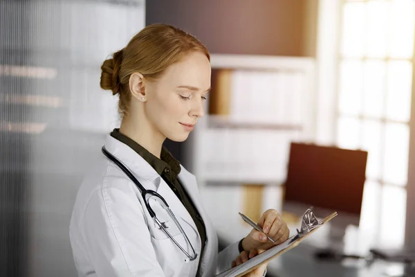 Friendly smiling female doctor using clipboard in sunny clinic. Portrait of friendly physician woman at work. Medicine concept