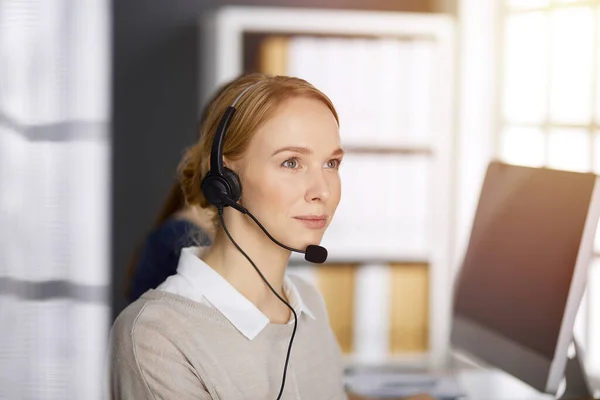 Amicale femme d'affaires caucasienne parlant par casque dans un bureau ensoleillé. Centre d'appels et divers groupes de personnes en affaires — Photo