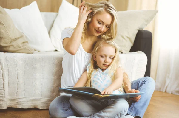 Bonne famille. Jeune mère blonde lisant un livre à sa jolie fille assise au sol. Concept de maternité — Photo