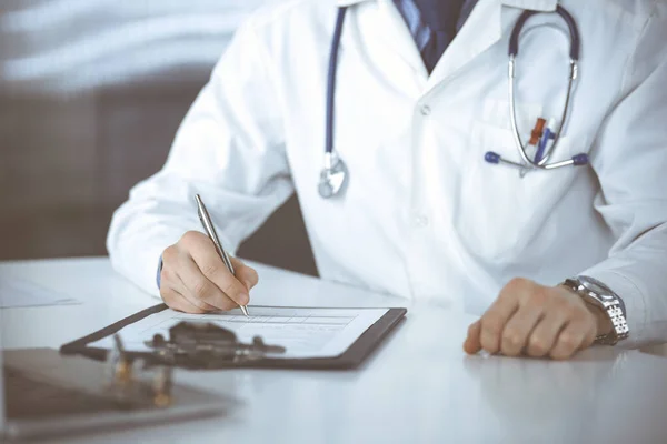 Okänd manlig läkare sitter och arbetar med urklipp av medicinhistoria rekord på kliniken på sin arbetsplats, närbild. Ung läkare på jobbet. Perfekt medicinsk service, medicin koncept — Stockfoto