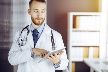 Dost canlısı sakallı doktor iş yerindeki güneşli klinikte tablet bilgisayar kullanıyor. Tıp konsepti