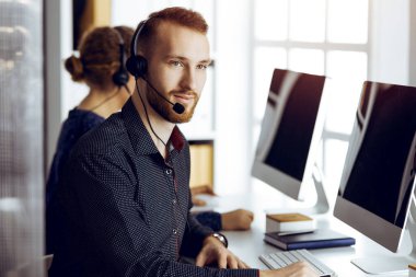 Modern güneşli ofiste otururken kulaklıkla konuşan sakallı iş adamı. Çağrı merkezinde çeşitli insanlar grubu