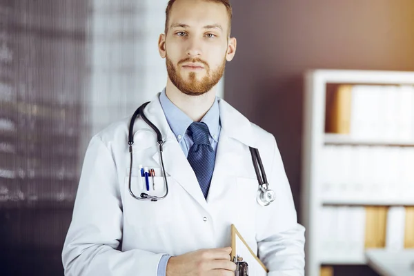 Friendly bearded doctor standing straight in sunny clinic. Portrait of physician. Medicine concept