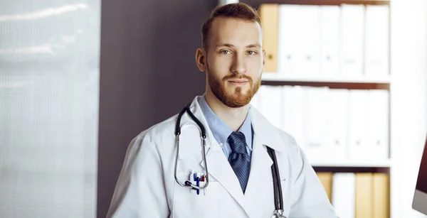 Amichevole medico barbuto seduto e scrivere negli appunti in clinica soleggiata. Concetto di medicina — Foto Stock