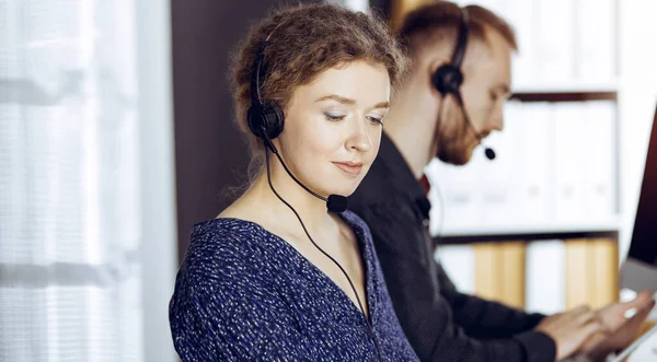 Femme d'affaires parlant par casque tout en étant assis avec un collègue à barbe rouge dans un bureau ensoleillé. Groupe de personnes diversifiées dans le centre d'appels — Photo