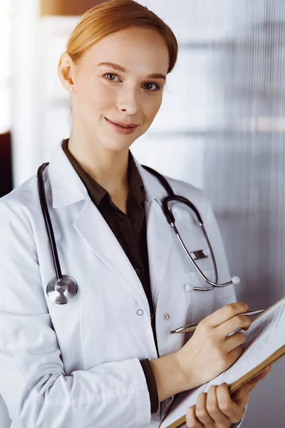 クリニックでクリップボードを使用して陽気な笑顔の女性医師。仕事中の優しい医師の女性の肖像画。病院で医療サービス。医学の概念 — ストック写真