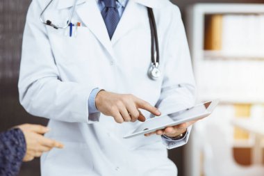 Bilinmeyen kızıl sakallı doktor ve hasta kadın güncel sağlık muayenesini güneşli klinikte otururken ve tablet bilgisayar kullanırken tartışıyorlar, yakın plan. Tıp konsepti