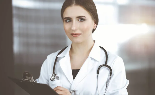 Amistosa doctora de pie y sujetando portapapeles en la clínica moderna. Retrato del alegre médico sonriente —  Fotos de Stock