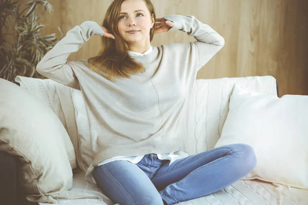 Glückliche brünette Frau legte die Hände hinter den Kopf und saß angelehnt auf der Couch, europäische weibliche Stretching und genießen faules Wochenende oder Urlaub im gemütlichen Wohnzimmer — Stockfoto