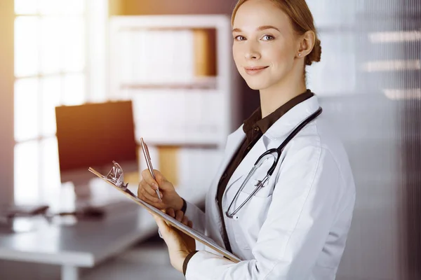 Allegro sorridente medico femminile utilizzando appunti in clinica. Ritratto di donna medico amichevole al lavoro. Servizio medico in ospedale. Concetto di medicina — Foto Stock